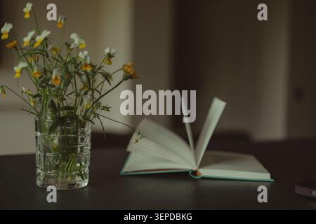 Stillleben mit einem offenen Buch und einem Blumenstrauß aus Wildblumen in einer Glasvase auf einem dunklen Tisch. Gemütliches Interieur, das Lesen, Freizeit, Quie symbolisiert Stockfoto
