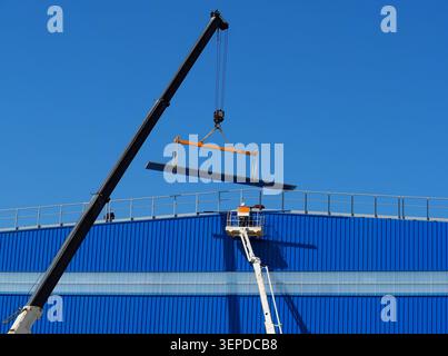 Cherry picher und Teleskopkran arbeiten an einer Fassade eines Industriegebäudes mit einem schweren Metallbalken. Nicht erkennbare Person, Stockfoto