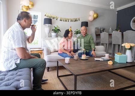Asiatische Familie feiert Geburtstag im Wohnzimmer mit einer älteren Frau, die Kerzen auf Kuchen bläst Stockfoto