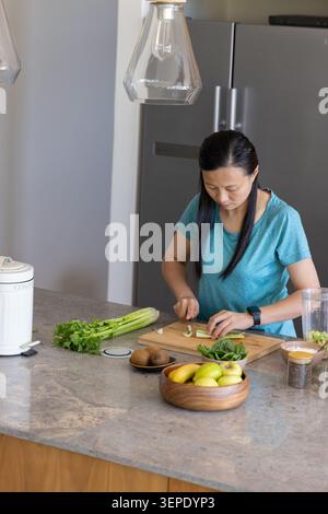 Asiatische Frau mittleren Alters, die Sellerie auf Schneidebrett in der Küche mit Mixbecher und Kopierraum hackt Stockfoto
