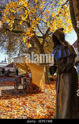 Bronze-Mönchsstatue mit Kapuze zwischen goldenen Herbstahornbäumen mit mittelalterlichem Holzwagen im Altstadtpark von Tallinn, Estland. Stockfoto
