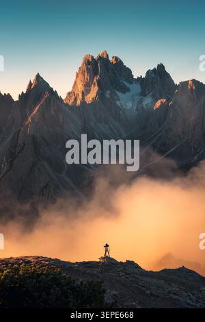 Spiegellose Kamera auf einem Stativ steht auf einem felsigen Kamm, während goldener Nebel durch den zerklüfteten Gipfel der Cadini di Misurina in den Dolomiten, Italien, in warmen Sonnenaufgang getaucht wird Stockfoto