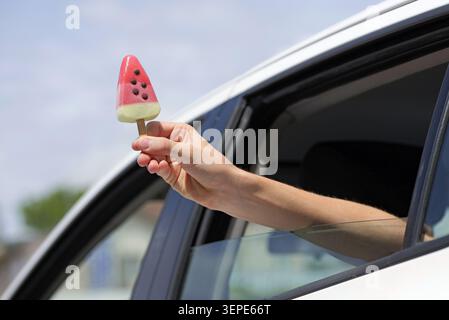 Die Hand zieht ein erfrischendes Wassermelonen-förmiges Eis aus einem beweglichen Autofenster, symbolisiert Freiheit, Reisen und einen gefrorenen Leckerbissen Stockfoto