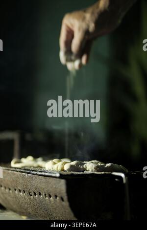 Mit der Hand Mehl auf kleine rohe Teigstücke streuen, die auf einem alten gusseisernen Grill kochen, wodurch Rauch mit einem dunklen, stimmungsvollen Hintergrund erzeugt wird, der trad betont Stockfoto