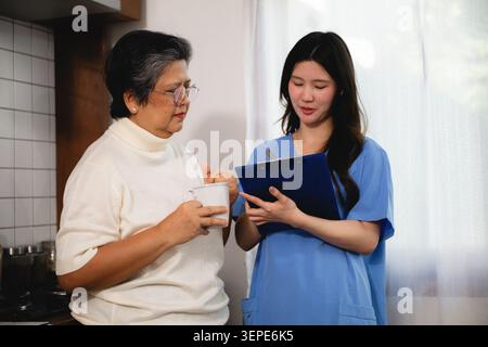 Home Health Care ältere Frau, ältere Frau hält Tasse in der Küche zu Hause, während Schwester in blauen Peelings hält Klemmbrett erklären, klären und empfehlen. Elde Stockfoto
