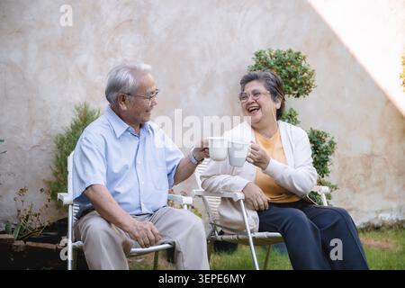 Ältere Männer und Frauen sitzen auf Terrassenstühlen. Sie teilen sich eine Tasse und lächeln warm. Das Sonnenlicht hebt ihre fröhlichen Ausdrucksformen hervor. Gartenpflanzen und eine Wand für Stockfoto