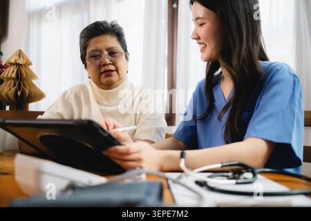 Ein medizinisches Pflegepersonal hält ein Tablet mit einem Patienten. Beide schauen zusammen auf den Bildschirm. Die jüngere Schwester lächelt sanft. Ein älterer Patient scheint aufmerksam zu sein Stockfoto