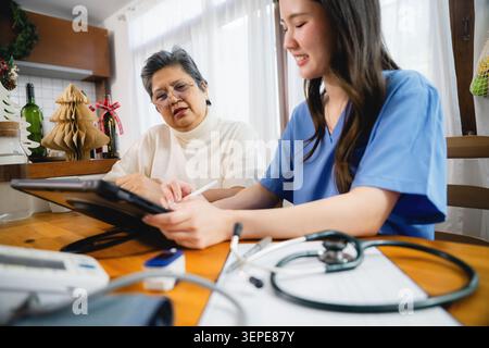 Ernsthafte ältere asiatische Frau sprechende Krankenschwester Beratung in gemütlichem Haus, professionelle Pflegekraft zuhört fokussierte reife Dame und Aufnahme auf Tablet mit Medizin Stockfoto