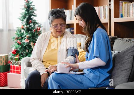 Die Krankenschwester überprüft den Blutdruck einer älteren Frau auf dem Sofa. Festliche Dekorationen füllen das gemütliche Wohnzimmer. Sie misst sorgfältig und zeigt professionelle Sorgfalt. Alt Stockfoto