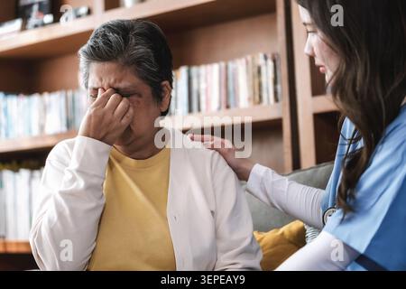Ältere Frau reibt sich die Schläfen vor Schmerzen. Ihre Schultern verspannen sich vor Unwohlsein. Die junge Pflegekraft legt ihr eine tröstende Hand auf die Schulter. Bücherregale Linie Stockfoto
