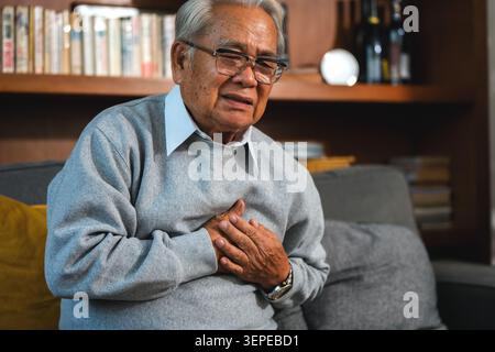 Der ältere Mann sitzt auf dem Sofa und die Hand auf der Brust. Angstzustände bei Herzinsuffizienz. Älterer pensionierter Großvater mit akutem Brustschmerz, Nahaufnahme sen Stockfoto