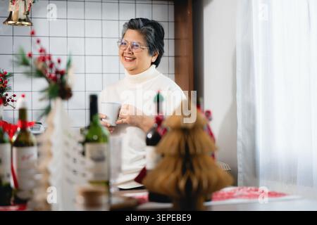 Glückliche ältere pensionierte Großmutter trinkt Tee weihnachtszeit, Brille lächelnd Winter Urlaub Haus Party, Lebensstil gesund alternde asiatische Großmutter Stockfoto