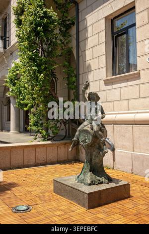Batumi, Georgia – 9. Juni 2026: Bronzeskulptur eines Jungen auf einem Weinfass nahe einer Mauer mit grünem Weinstock. Stockfoto