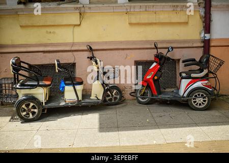 Batumi, Georgia - 9. Juni 2026: Ein langes beigefarbenes Dreirad und ein rot-weißer Single Roller mit Gepäckkörben, die auf einem Bürgersteig geparkt sind. Stockfoto