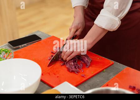 Nahaufnahme einer Frau, die rohes Fleisch in kleine Stücke auf rotem Brett zum Kochen schneidet. Stockfoto