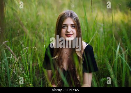 Junge Frau mit fließenden braunen Haaren, die friedlich zwischen hohem grünem Gras kniet, in elegantem schwarzen Kleid Stockfoto