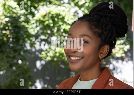 Erwachsene afrikanische Frau, die im grünen Hinterhof steht und lächelt, Rostjacke und Ohrring trägt Stockfoto