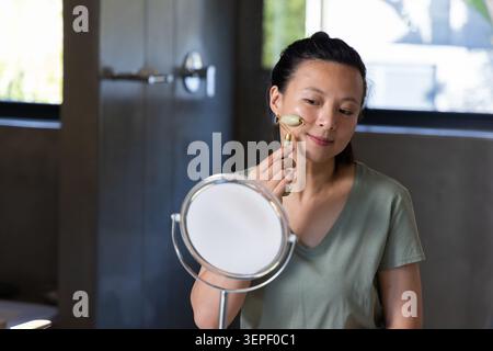 Asiatische Frau in Salbei-T-Shirt, die Steinrolle auf die Wange am Badezimmertisch mit Tischspiegel aufträgt Stockfoto