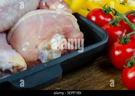 Rohe Hähnchenschenkel und Drumsticks mit frischen Tomaten und Paprika in schwarzer Pfanne auf Holztisch, gesunde Zutaten. Nahaufnahme. Stockfoto