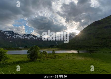 Dramatische Berglandschaft mit einem ruhigen See, saftig grünen Wiesen und schneebedeckten Gipfeln, die von der untergehenden Sonne beleuchtet werden. Sonnenstrahlen brechen durch Stockfoto