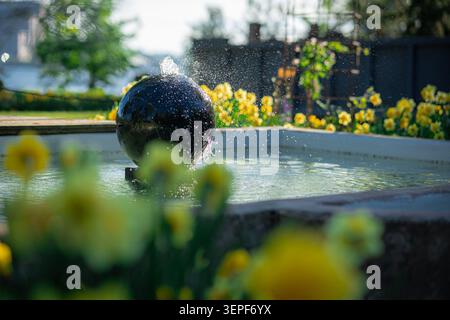 Dunkler, kugelförmiger Steinbrunnen, der Wasser in einem sonnigen Garten sprüht. Die Wassertropfen funkeln im Sonnenlicht, umgeben von einem wunderschönen Blütenfleck Stockfoto