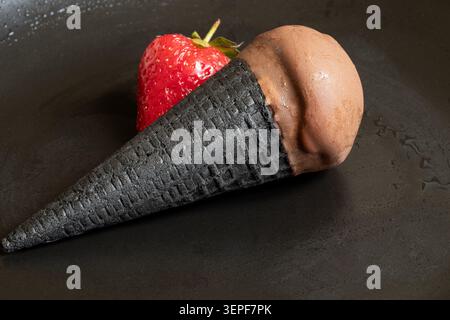 Schokoladeneis in einem schwarzen Waffelkegel und eine einzelne Erdbeere auf einem schwarzen Teller, ein erfrischendes kaltes Dessert mit Schokoladenglasur Stockfoto