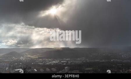 Dramatischer Blick auf Sonnenstrahlen, die durch dunkle Sturmwolken über einer europäischen Stadt, Tal, Hügel und entfernte Regenschauer brechen Stockfoto