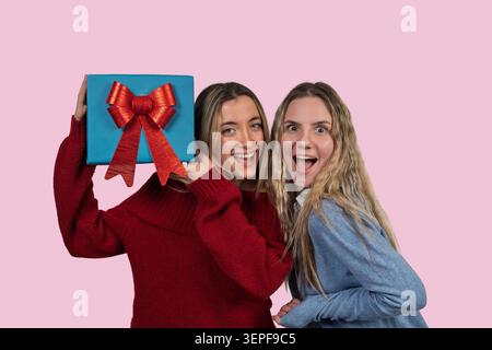 Zwei aufgeregte Freunde teilen einen freudigen Moment mit einem Überraschungsgeschenk auf rosa Hintergrund Stockfoto
