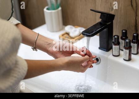 Hände waschen Seife unter schwarzem Badezimmerhahn. Nahaufnahme der Hände, die einen Seifenstab unter fließendem Wasser in einem weißen Waschbecken mit Matte waschen Stockfoto