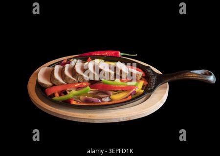 Gebratene Schweinemedaillons mit gegrilltem Gemüse in einer heißen Pfanne. Das Essen ist auf schwarzem Hintergrund isoliert. Stockfoto