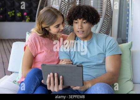 Ein Paar sitzt auf einem Sofa im Freien auf dem überdachten Balkon, hält ein Tablet in der Hand, trägt T-Shirts und Jeans Stockfoto