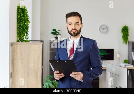 Selbstbewusster Geschäftsmann im Anzug hält Klemmbrett für Firmenporträt Stockfoto