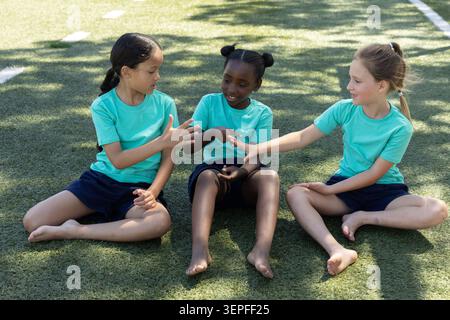 Verschiedene Schulmädchen sitzen barfuß auf dem Rasen in passenden blauen Hemden marineblauen Shorts und spielen Handspiele Stockfoto