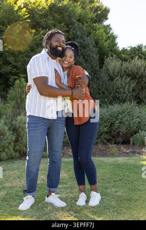Afroamerikanisches Paar umarmt sich auf dem Park Rasen in mehrschichtigen Hemden weißen Sneakers Armband Linse Flare Stockfoto