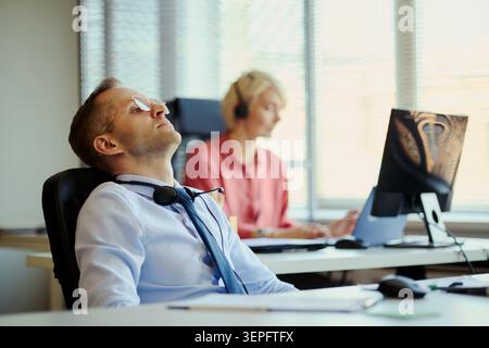 Junger erwachsener kaukasischer Mann, der während der Callcenter-Schicht ruht, während eine Kollegin im Büro am Computer arbeitet. Ideal für Kundensupport, Burnout, Stress-Kampagnen am Arbeitsplatz Stockfoto