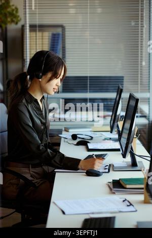 Vertikale Aufnahme einer jungen, asiatischen Frau, die im Callcenter arbeitet, ein Headset trägt, Notizen macht und den Workflow des Kundensupports im Teambüro überwacht. Für Service Business Marketing Stockfoto