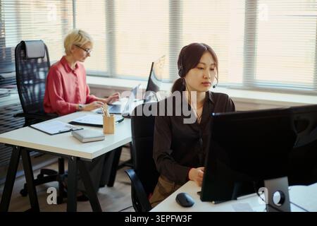Asiatische junge Erwachsene Frau mit Headset, die im Callcenter am Computer arbeitet, kaukasische Frau mittleren Alters, die in der Nähe schreibt. Ideal für Kundensupport, CRM und Teamwork-Marketing Stockfoto