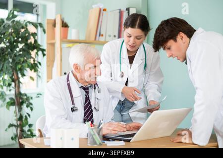 Professioneller älterer Mann Arzt und zwei junge männliche und weibliche Praktikanten oder Krankenschwestern, die sprechen, mit Laptop arbeiten und Patientenpapiere im Arzt überprüfen Stockfoto
