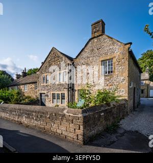 Breiter Panoramablick auf das historische aus Stein gebaute Hawksworth Cottage hinter einer niedrigen Steinmauer an einer Dorfstraße Ecke in Eyam Stockfoto
