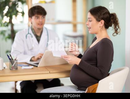 Erfahrener junger männlicher Arzt in weißem medizinischen Mantel und Stethoskop, der schwangere Patientin, die am Tisch in der medizinischen Praxis im Krankenhaus sitzt, berät Stockfoto