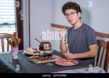 Junger Mann, der an einem Tisch sitzt und burgundisches Fondue mit kochendem Öl genießt, Spieße und Fleisch isst, ein Glas Rotwein in der Nähe in einer gemütlichen Wohnung dini Stockfoto