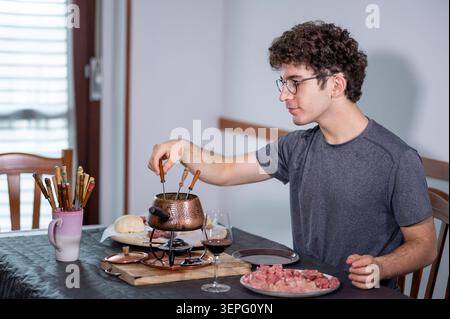 Junger Mann an einem Esstisch, der Fleischwürfel in einem traditionellen Kupferfonduetopf mit kochendem Öl kocht, das Essen und ein Glas Rotwein in einem genießt Stockfoto