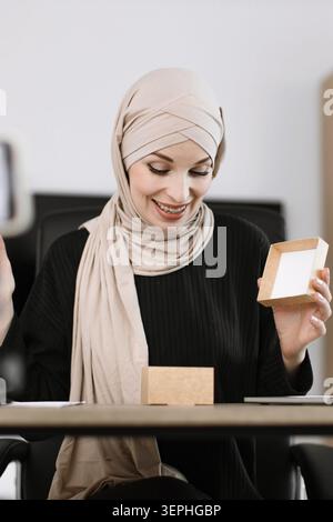 Eine lächelnde Frau mit Hijab öffnet einen kleinen Karton und enthüllt seinen Inhalt Stockfoto