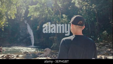 Ein Mann steht und blickt auf den tropischen Wasserfall im üppigen grünen Dschungel-Wald. Rückansicht der Reisenden, die die Natur während des sonnigen Tages erkunden. Abenteuer und draußen Stockfoto
