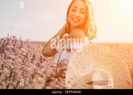 Lavendelfeldfrau lächelnde junge Frau genießt die goldene Stunde Sonnenuntergang auf einer blühenden Wiese, während sie einen Strohhut hält Stockfoto