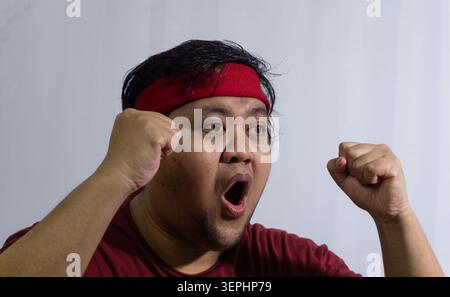 Überraschte indonesische Sportfans, die überraschend beim Anschauen eines Spiels keuchten und mit rotem Hemd und h seine Fäuste in intensiver Begeisterung geballt haben Stockfoto