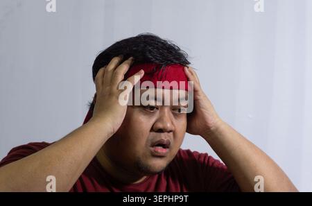 Der ängstliche indonesische Sportfan hält sich nach einem verpassten Tor verzweifelt den Kopf und zeigt einen gestressten und besorgten Ausdruck in einem roten Hemd und Kopf Stockfoto
