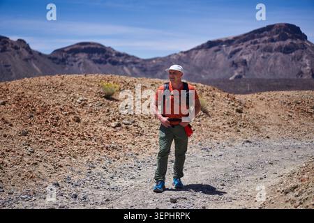 Ein erwachsener männlicher Wanderer mit Rucksack und Mütze steht auf dem vulkanischen Pfad von Montana Blanca mit dem Las-Cañadas-Kamm im Hintergrund, dem Teide Nat Stockfoto