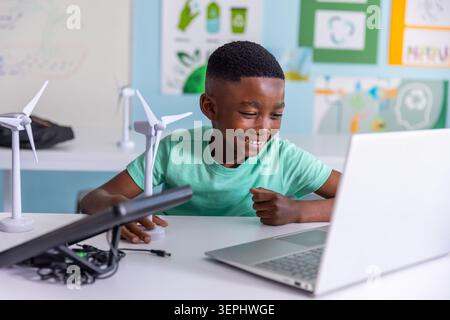 Afroamerikanischer Junge in grünem T-Shirt, der am Schreibtisch sitzt und mit Laptop- und Windturbinenmodellen arbeitet Stockfoto