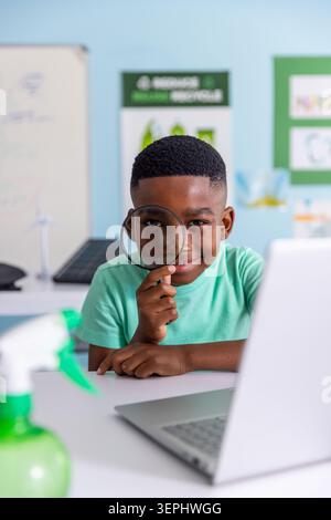 Afroamerikanischer Schuljunge mit Lupe, der in der Schule auf Laptop-Wind- und Solarmodelle blickt Stockfoto